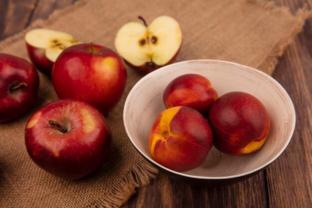 Вид сверху свежих персиков на миске с яблоками, изолированными на мешковине на деревянной стене