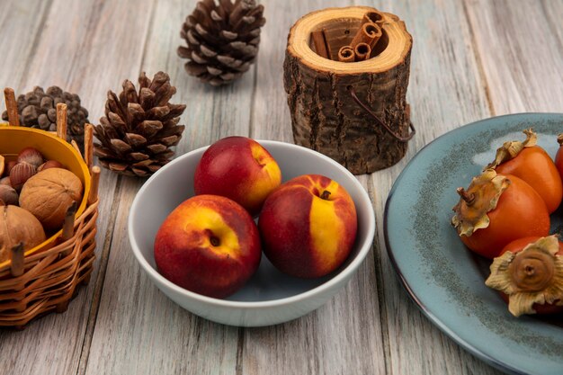 Top view of fresh peaches on a bowl with persimmons on a plate with cinnamon sticks on a grey wooden surface