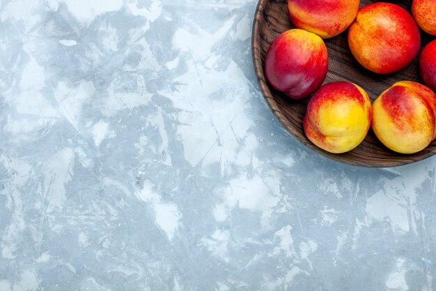 Вид сверху свежие персики, спелые и вкусные фрукты внутри коричневой тарелки на светло-белом столе