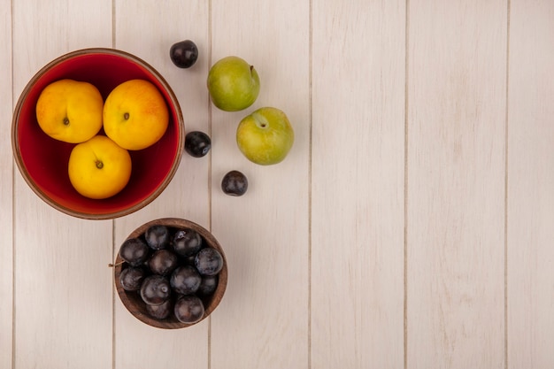 Free photo top view of fresh peaches on a red bowl with sloes on a wooden bowl with green cherry plums isolated on a wooden background with copy space