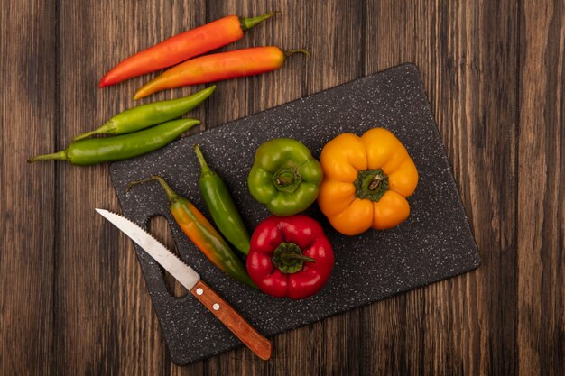 Top view of fresh peppers on a black kitchen board with knife with peppers isolated on a wooden wall