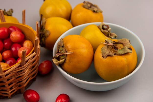 Top view of fresh persimmon fruits on a bowl with cornelian fruits on a bucket on a grey surface