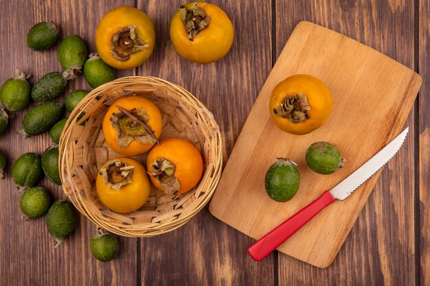 Top view of fresh persimmon fruits on a bucket with feijoas and persimmon on a wooden kitchen board with knife on a wooden surface