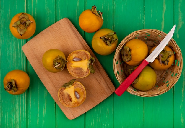Вид сверху свежих фруктов хурмы на ведре с ножом с разрезанными пополам плодами хурмы на деревянной кухонной доске на зеленом деревянном столе