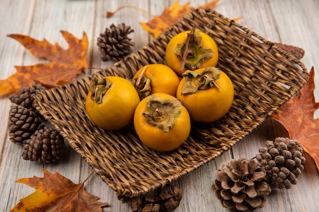 Top view of fresh persimmon fruits on a wicker tray with leaves on a grey wooden table