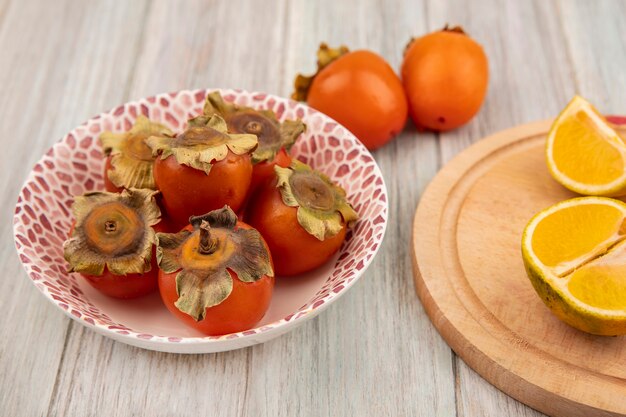 Top view of fresh persimmons on a bowl with tangerines on a wooden kitchen board on a grey wooden wall