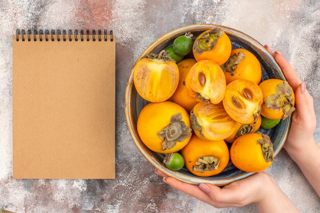 Top view fresh persimmons feykhoas in a bowl in female hand a notebook on nude background