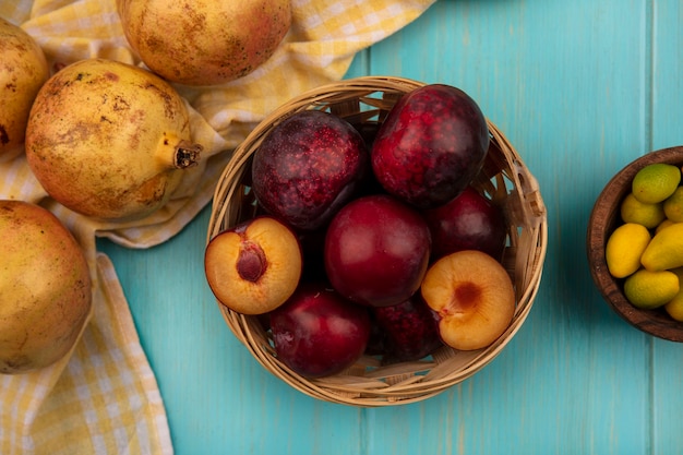 Top view of fresh pluots on a bucket with yellow pomegranates isolated on a yellow checked cloth with kinkans on a wooden bowl on a blue wooden wall