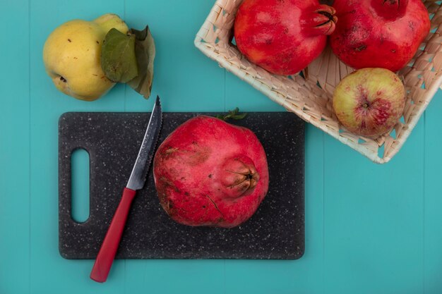 Top view of fresh pomegranate on a black kitchen board with knife with a bucket of fruits on a blue background