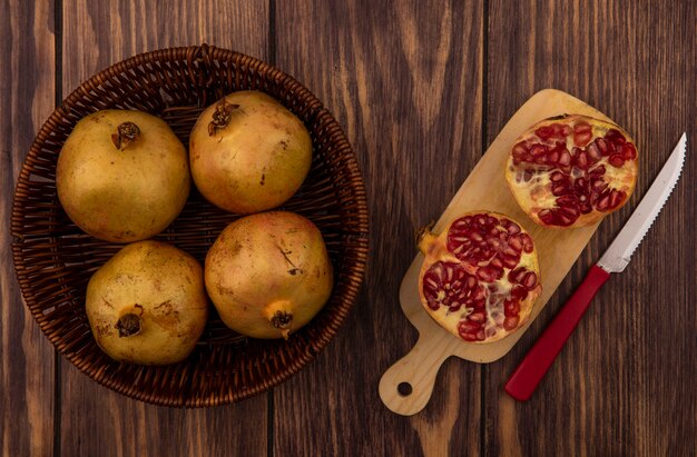 Top view of fresh pomegranates on a bucket with halved pomegranates on a wooden kitchen board with knife on a wooden wall