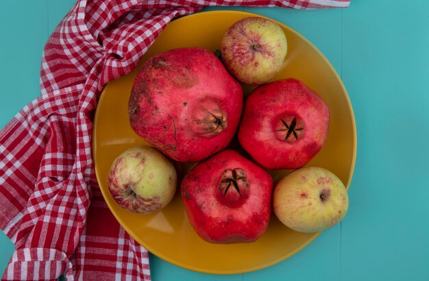 Top view of fresh pomegranates on a yellow plate with apples on a checked plate on a blue background