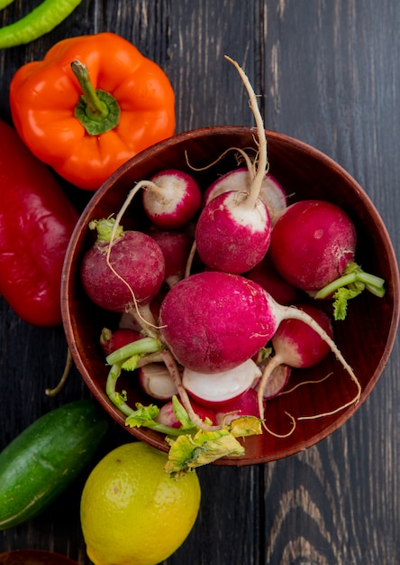 Top view of fresh radish in a wood bowl and fresh colorful bell peppers cucumbers and lemon on dark wood