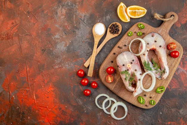 Top view of fresh raw fishes and pepper onion greens tomatoes on wooden cutting board on the left side on mixed color surface