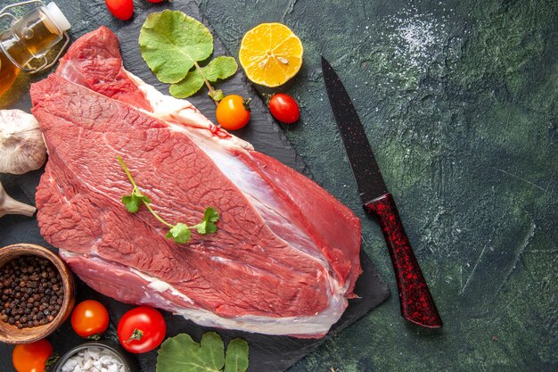 Top view of fresh raw red meat on black tray pepper vegetables fallen oil bottle knife on dark color background