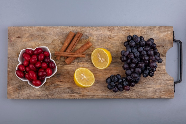 Top view of fresh red cornel berries on a bowl with lemon cinnamon sticks and grapes on a wooden kitchen board on a grey background