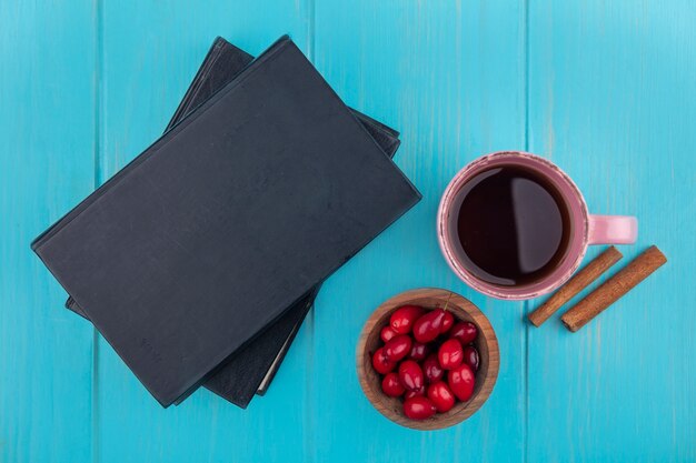 Top view of fresh red cornel berries on a wooden bowl with a cup of tea and cinnamon sticks on a blue wooden background