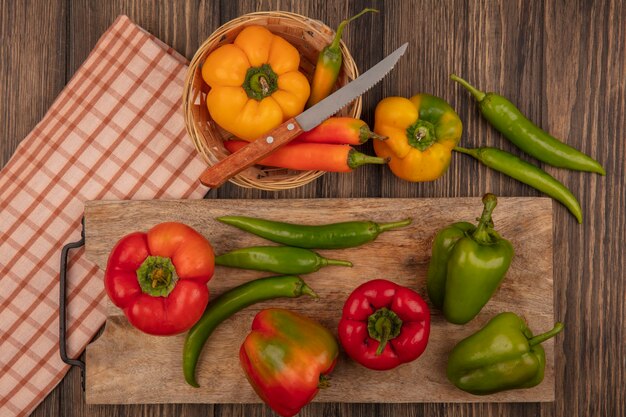 Top view of fresh red and green peppers on a wooden kitchen board with yellow peppers on a bucket with knife on a checked cloth on a wooden wall