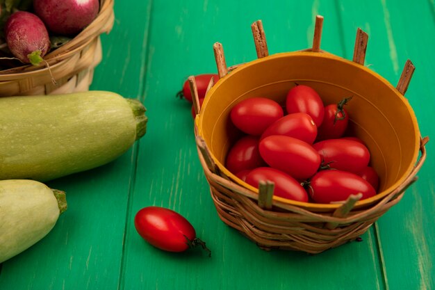 Top view of fresh red plum tomatoes on a bucket with radishes on a bucket with zucchinis isolated on a green wooden wall