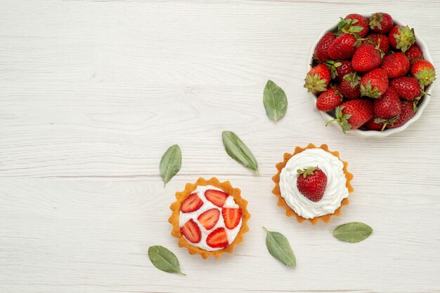 Вид сверху свежей красной клубники, спелых и вкусных ягод внутри белой тарелки с тортами на свету, ягодами красного цвета