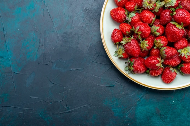 Вид сверху свежей красной клубники, спелых фруктов, ягод внутри тарелки на темно-синем столе, ягодных фруктов, спелого лета