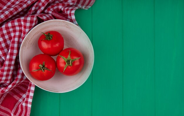 Вид сверху свежих и красных помидоров на миске на красной клетчатой ткани на зеленом деревянном фоне с копией пространства