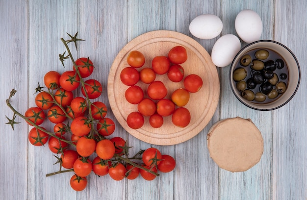 Free photo top view of fresh red vine tomatoes on a bowl with tomatoes isolated on a wooden kitchen board with olives on a bowl and eggs on a grey wooden background