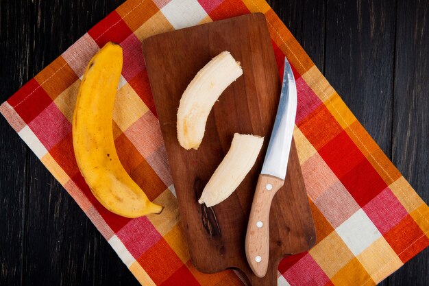 Top view of fresh ripe banana fruit and peeled sliced banana on a wooden cutting board with kitchen knife on rustic