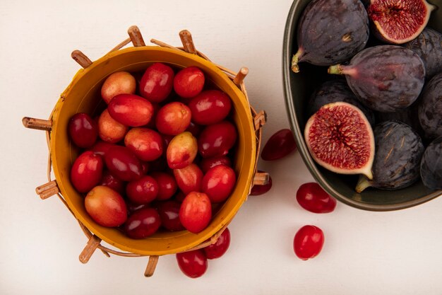 Top view of fresh ripe black figs on a bowl with cornelian cherries on a bucket on a white wall