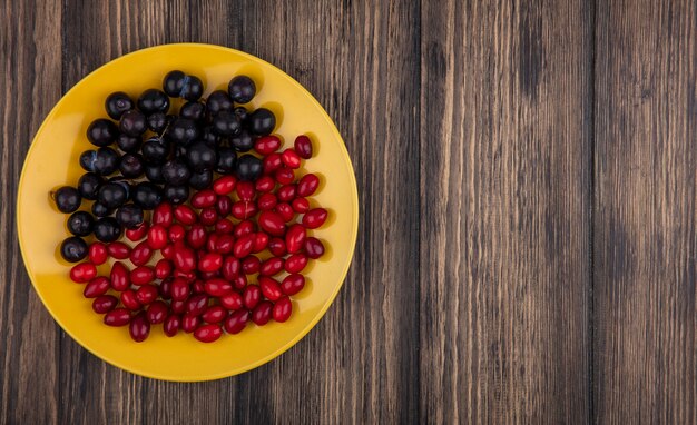 Top view of fresh ripe cornel berries with blackthorns on a yellow plate on a wooden background with copy space