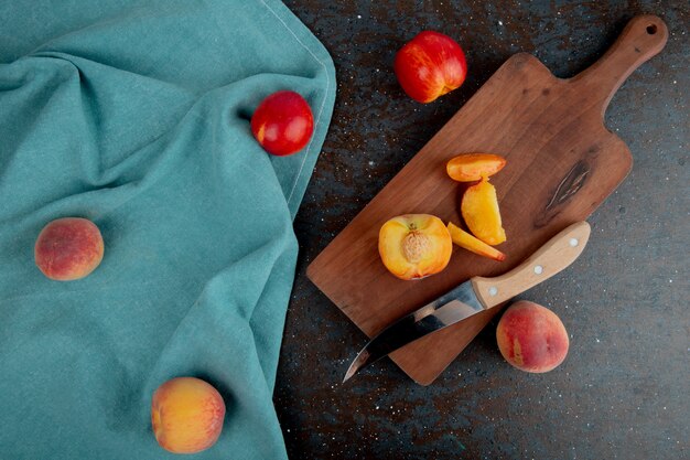 Top view of fresh ripe nectarine and slices with kitchen knife on a wooden cutting board on blue fabric on black