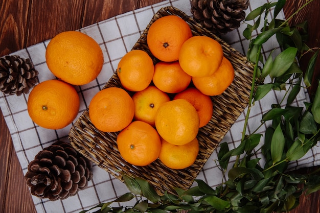 top view of fresh ripe oranges on a wicker tray and cones on plaid tablecloth on wooden surface