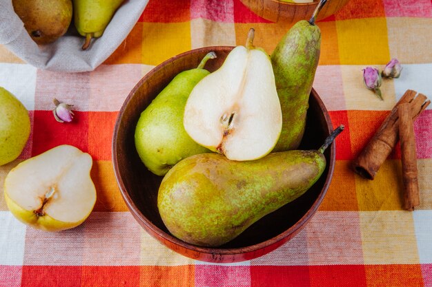 Top view of fresh ripe pears and a half in a wooden bowl and cinnamon sticks on plaid