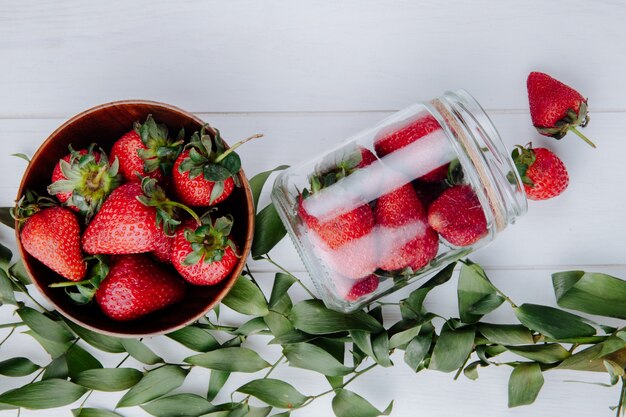 top view of fresh ripe strawberries in a wooden bowl and strawberries scattered from a glass jar and green leaves on white wooden table