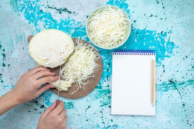 Top view of fresh sliced cabbages with half whole vegetable getting cut and notepad on bright-blue desk, vegetable food meal snack healthy salad