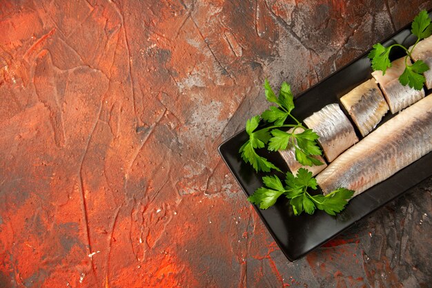 Top view fresh sliced fish with greens and lemon pieces inside black pan on dark background