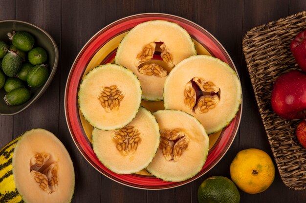 Top view of fresh slices of cantaloupe melon on a plate with feijoas on a bowl with apples on a wicker tray on a wooden background