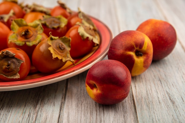 Free photo top view of fresh soft and juicy persimmons on a plate with peaches isolated on a grey wooden wall