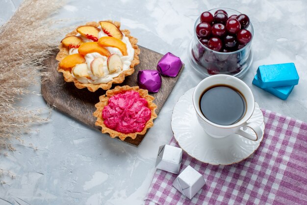Вид сверху свежей вишни в маленькой стеклянной чашке со сливочными тортами и чаем на белом столе, фруктовый кисло-витаминный сладкий