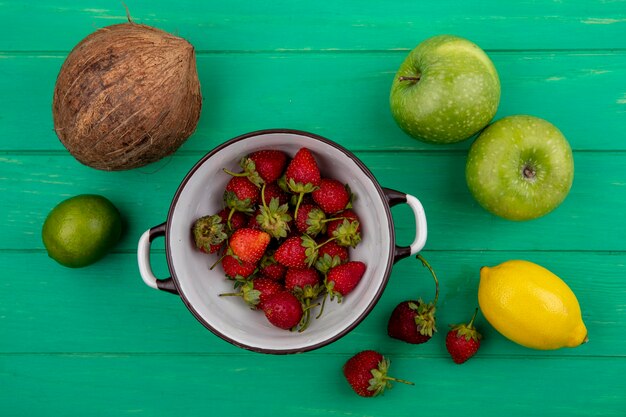 Top view of fresh strawberries on a bowl with fruits such as applelemonlimecoconut on a green wooden background