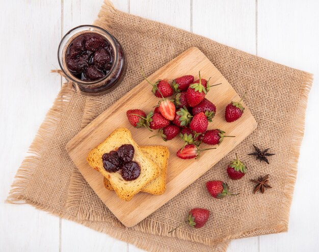 Top view of fresh strawberries on a wooden kitchen board on a sack cloth with strawberry jam on white background