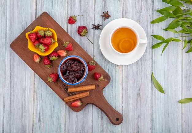 Top view of fresh strawberries on a yellow bowl on a wooden kitchen board with a strawberry jam with cinnamon sticks with a cup of tea with leaves on a grey wooden background