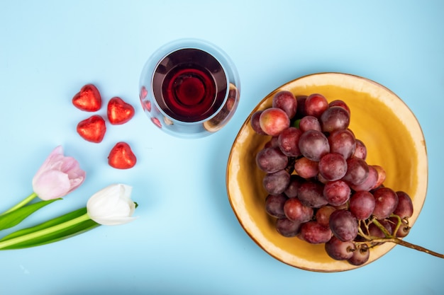 Top view of fresh sweet grape in a bowl with white and pink color tulips, a glass of wine and heart shaped chocolate candies in red foil scattered on blue table