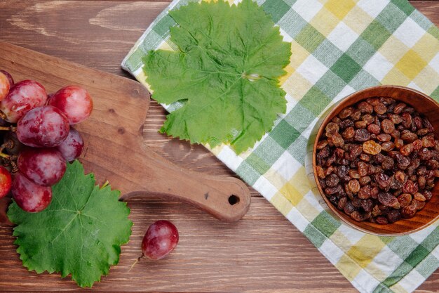 Top view of fresh sweet grape on a wooden cutting board , green grape leaves and raisins in a bowl on plaid fabric on wooden table