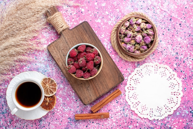 Top view of fresh tasty raspberries inside white plate with tea and cinnamon on pink surface