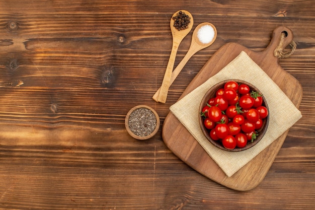 Top view of fresh tomatoes on a brown bowl on cutting board and spices in spoons pepper on wooden board