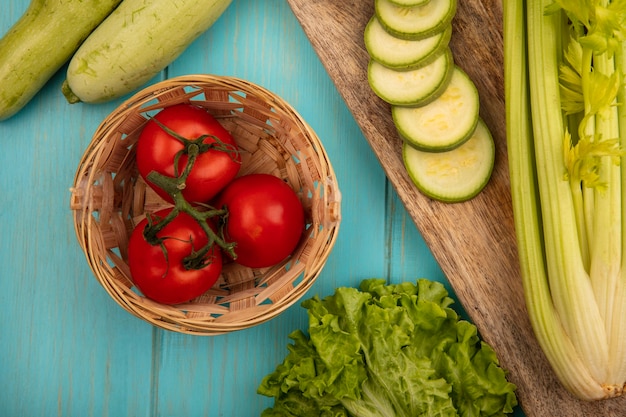 Top view of fresh tomatoes on a bucket with chopped zucchinis on a wooden kitchen board with celery with lettuce isolated on a blue wooden surface