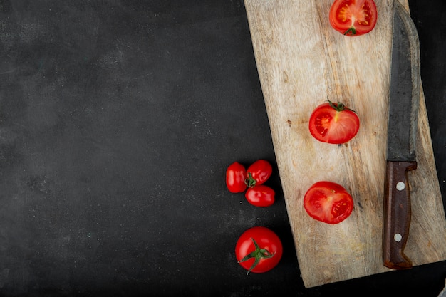 Top view of fresh tomatoes and halves on wooden chopping board next to a kitchen knife on black background