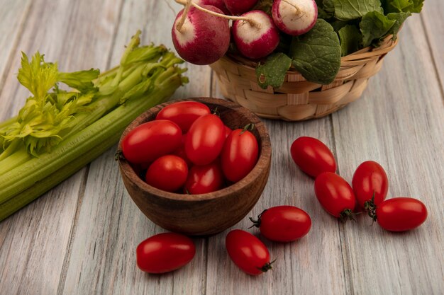 Top view of fresh tomatoes on a wooden bowl with radishes on a bucket with tomatoes and celery isolated on a grey wooden wall