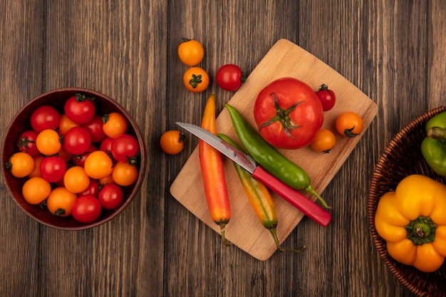 Top view of fresh tomatoes on a wooden kitchen board with long peppers with knife with cherry tomatoes on a wooden bowl and bell peppers on a bucket on a wooden surface