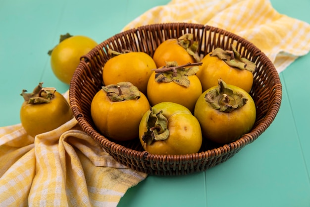 Top view of fresh unripe persimmon fruits on a bucket on a yellow checked cloth on a blue wooden table